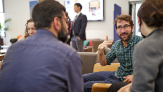 Paulo Savaget sitting with a group of people gesticulating and talking, wearing a green check shirt. 
