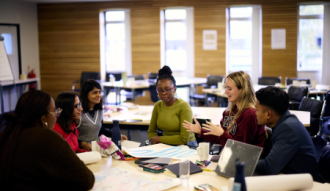Picture of 6 students sitting around a table in discussion with papers and pens in the middle