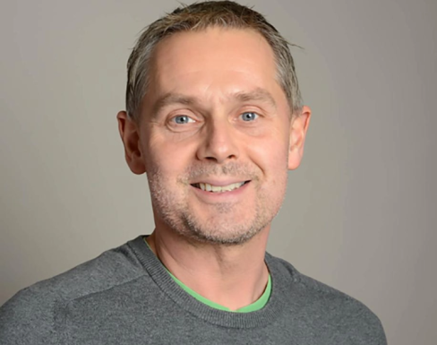 Alex Nicholls standing in front of a grey background and smiling.