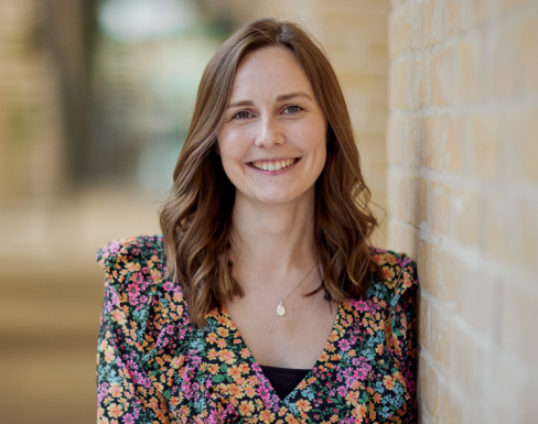 Lydia Allard standing in the SBS hallway wearing a floral top and smiling. 