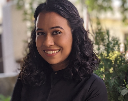 Simeen Kaleem standing in front of a leafy background wearing a dark suit and smiling.