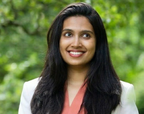 Sugandha Srivastav smiling standing in front a leafy and bright green background. 