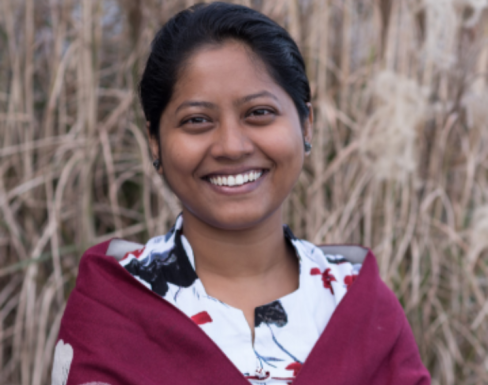 Anjali Sarker in front of a field smiling wearing a white patterned blouse with a red shawl.