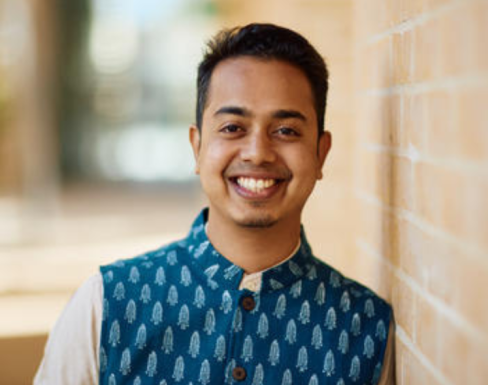 Avinash Kothuri smiling and standing in the SBS hallway wearing a blue patterned button-up and pale shirt.