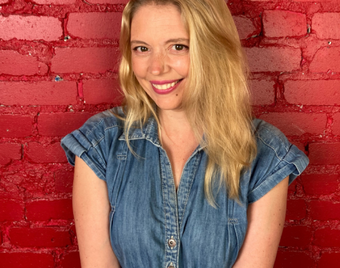 Charmian Love smiling in front of a red painted brick wall wearing a denim jumpsuit.