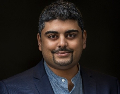 Darshan Sanghrajka standing against a black background wearing a blue button up and navy blue blazer.