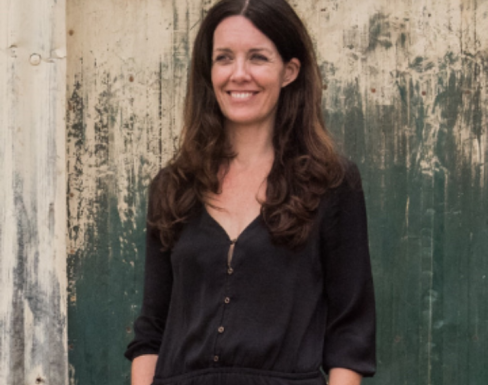Emma Caddy standing in front of a wooden panel in a dark grey blouse, smiling.