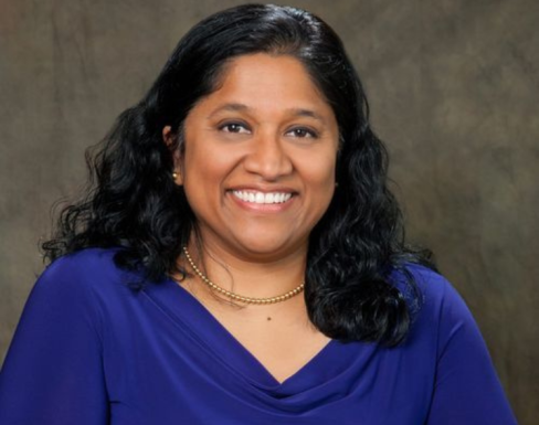 Esther Benjamin smiling in a royal blue dress and standing against a grey background.