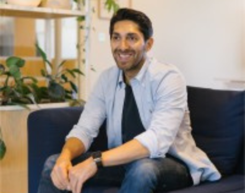 Faisal Butt sitting on a blue sofa with plants behind. He is smiling and wearing a pale blue shirt and jeans. 