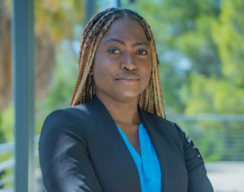 Folasade Ayoola is pictured standing with her arms crossed in front of a blurred natural background, wearing a blue shirt and black blazer.