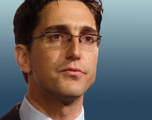 François Bonnici  in front of a blue background wearing a beige suit and tie.