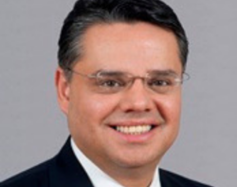 Henry Gonzalez standing in front of a grey background wearing a suit and red tie.