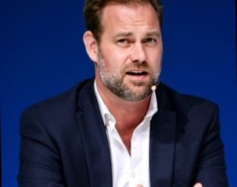 Jesse Moore sitting in a black blazer and white shirt talking in front of a blue background.