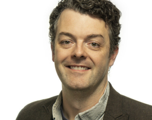 Kevin Warner smiling and wearing a brown suit. 