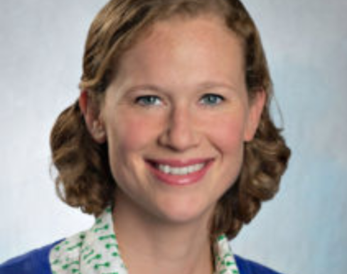 Lara Vogel smiling in front of a pale blue background wearing a blue cardigan and white shirt.