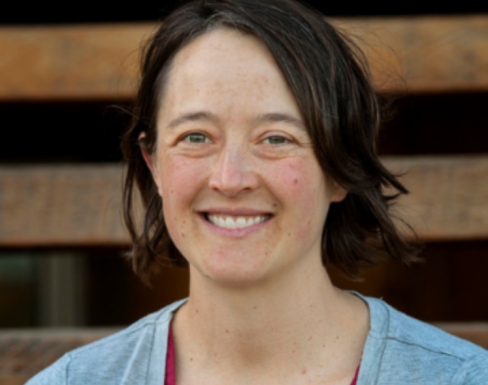 Lindsay Miller standing and smiling in front of wooden panels wearing a blue cardigan.