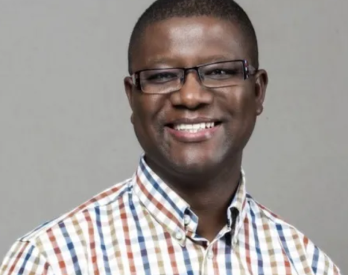 Martin Kalungu-Banda smiling in front of a grey background wearing a white, blue, beige and dark red checked shirt. 