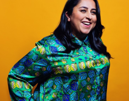 Maryam Pasha standing in front of a bright mustard yellow background, laughing and wearing a bright blue and emerald patterned shirt.
