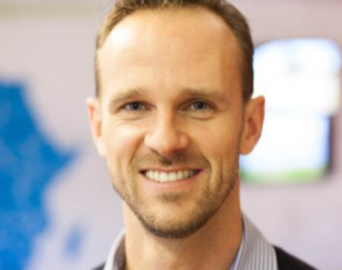 Mike Quinn smiling in front of a blurred background wearing a blue shirt.