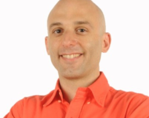 Nick Andreou smiling in front of a white background wearing a bright orange shirt.