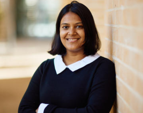 Nitya Nangalia leaning against a brick wall in the SBS courtyard, smiling and wearing a black sweater with white shirt. 