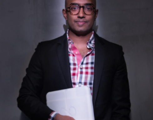 Rangan Srikhanta is pictured standing in a spotlight holding a laptop, wearing a blue and red checked shirt.