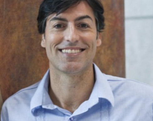 Richard Webb smiling in front of a wooden panel in a light blue shirt.