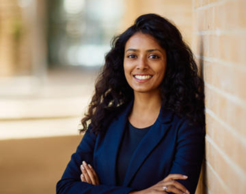 Shruti Jain leaning against a brick wall in the Said Business School courtyard, smiling and wearing a black blouse and navy blue blazer.