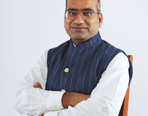 Sujeet Kumar sitting with his arms crossed in front of a white background wearing a pin stripe sleeveless button-up and white shirt.  