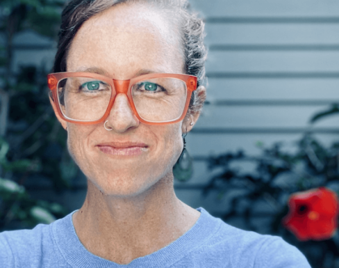 Sydney Schaef wearing bright red glasses and a blue top and smiling. 
