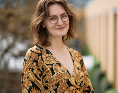 Menna Clarke standing in SBS wearing a yellow shirt and smiling. 
