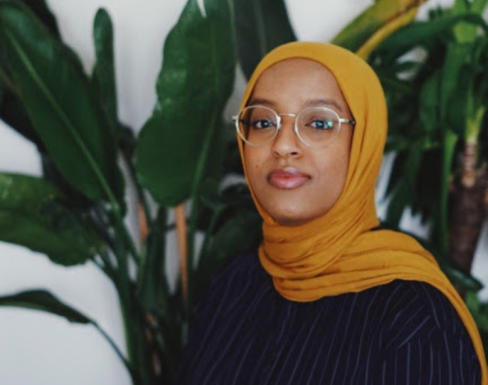 Izzy Mohammed standing in front of a green plant wearing a black top. 
