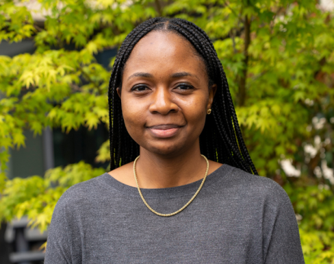 Aisha Raheem wearing a grey top in front of a green bush. 