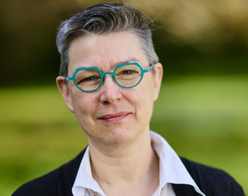 Close up headshot of Kathryn wearing a white shirt, black cardigan and circular turquoise-framed glasses. 