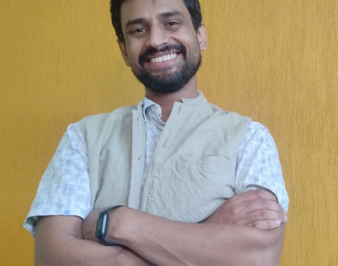Saransh Vaswani is standing in front of a yellow wall with his arm crossed. He is smiling and wearing a white top. 