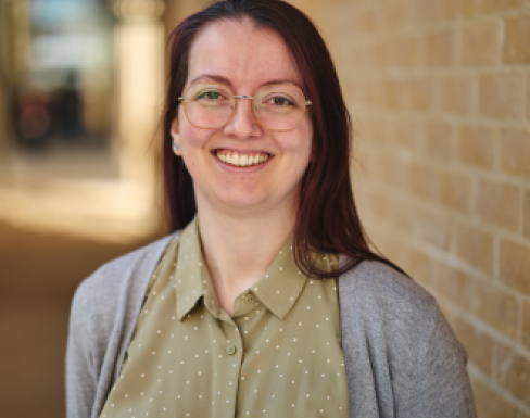 Close up shot of Alycia in front of a brick wall. She is smiling and wearing glasses, and a khaki yellow polka dot shirt with a grey cardigan. 