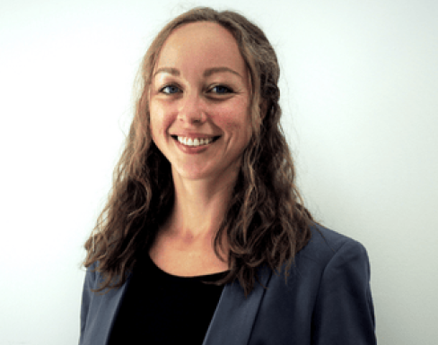Headshot from the shoulders up of Astrid smiling in front of a white background. She is wearing a blue grey suit jacket with a black top. 
