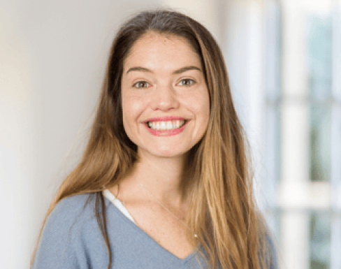 Headshot from shoulders up of Hayley grinning, in a well lit room. She is wearing a grey blue v neck jumper. 