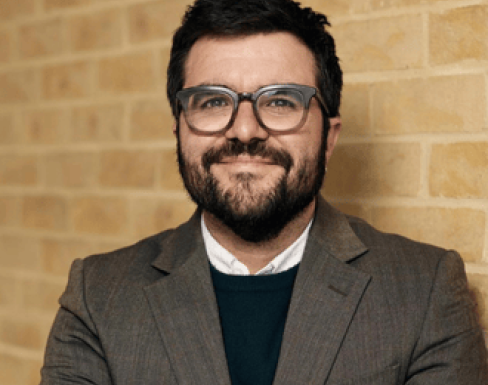 Headshot from shoulders up of Ignacio smiling in front of a yellow brick wall. He wears thick rimmed glasses, and a brown suit jacket with white shirt and dark brown jumper underneath. 