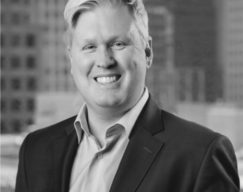 Gary is wearing a suit in front of skyscrapers smiling. 