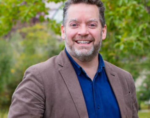 James Stauch is smiling in front of greenery, wearing a brown jacket and a blue shirt. 