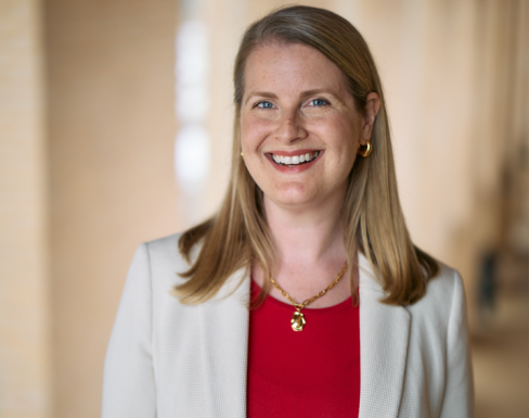 Mary is wearing a cream blazer and a red top. She is standing in a hallway, smiling. 