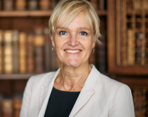 Mette is wearing a cream jacket and is smiling in front of a bookcase.