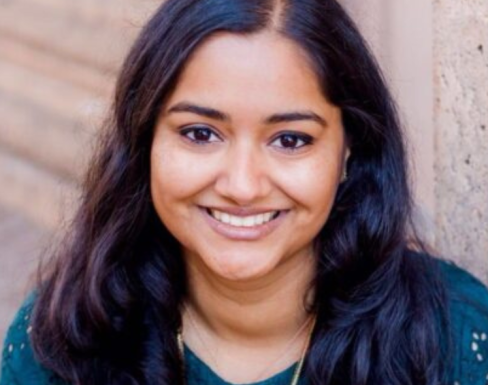 Shivani in standing in front of a brick wall and smiling wearing a blue top. 