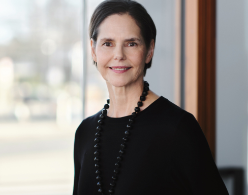 Johanna is wearing a black top and a large necklace in front of a bright window.