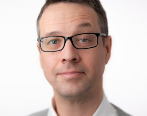 Headshot of Howard. He wears glasses, has short dark hair and is wearing a white shirt with grey blazer. 