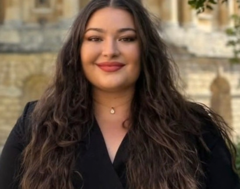 Headshot of Jasmine with an Oxford building in the background. She has long brown hair and is wearing a black top.