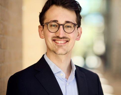 Headshot of Lukas in a warmly lit hallway. He wears round glasses, is smiling and wears a black blazer and pale blue shirt. 