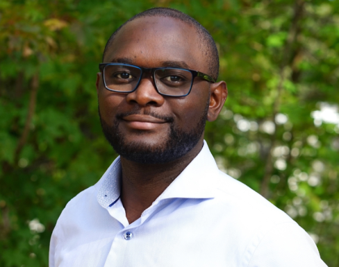 Headshot of Maganizo in front of a green hedge. He wears glasses, has very short hair and is wearing a white shirt. 