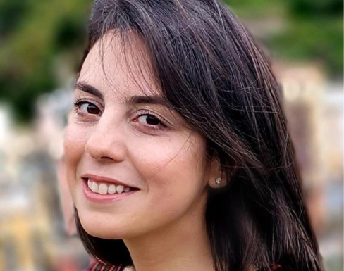 Heasdshot of Vira in front of a green/urban blurred background. She has long dark hair and is wearing a black and red checkered top. 